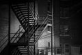 Black-and-white New York scene with a metal fire escape casting angular shadows on brick walls and shoes hanging from overhead wires.