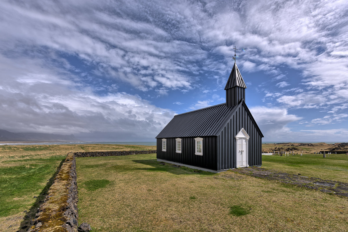 Black wooden church with white door and windows in Budir, Iceland, surrounded by grass and a low stone wall beneath textured clouds.