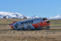 Weathered red-and-silver aircraft wreck on open ground with snow-covered mountains and a blue sky in Iceland.