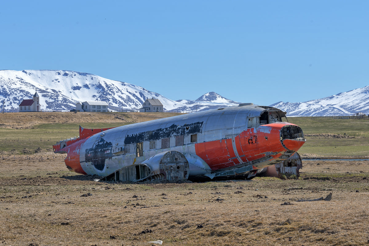 Weathered red-and-silver aircraft wreck on open ground with snow-covered mountains and a blue sky in Iceland.