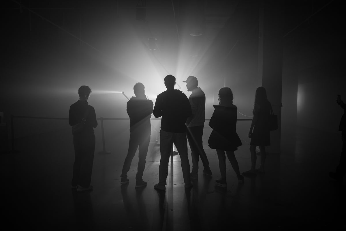 Black-and-white photograph of silhouetted people standing in a dim interior against a bright, hazy light, with reflections on the floor.